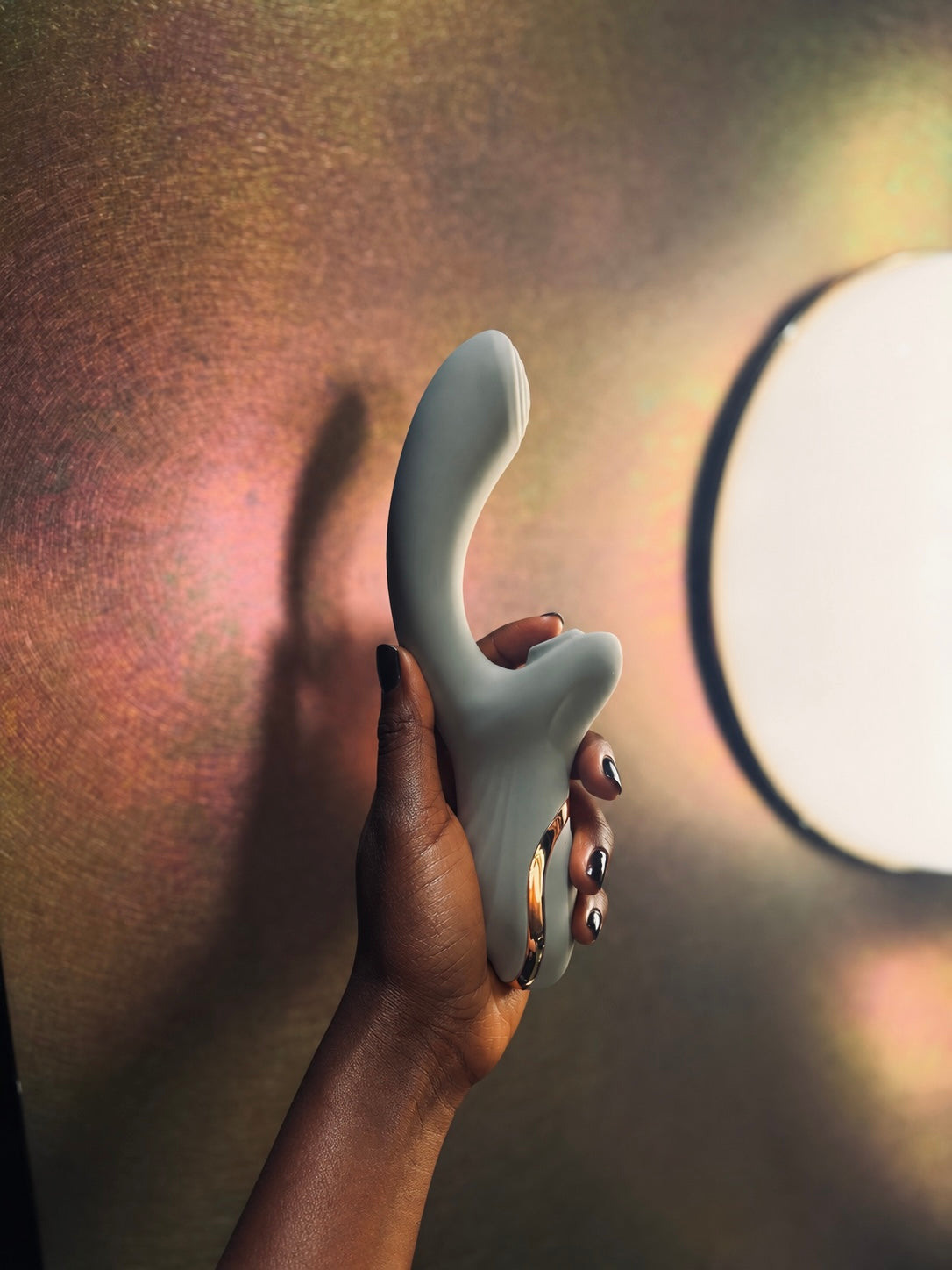 A hand holding The Duet, a dual-stimulation suction vibrator, against warm iridescent light